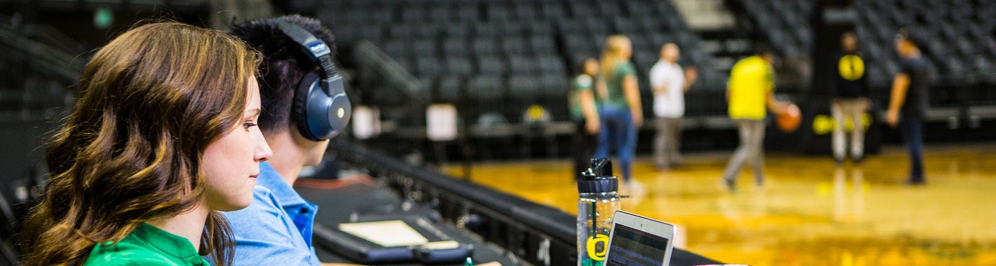 Two students sitting on the sidelines of a basketball court using laptops