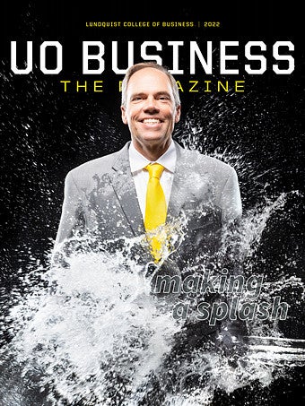A man wearing a gray suit with a yellow tie is standing against a black backdrop and being splashed with water.
