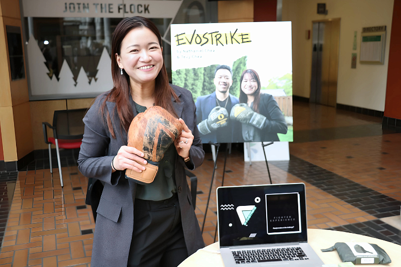 A student is holding up a prototype of her boxing glove for her startup "Evostrike" and standing in front of a poster with her project details.