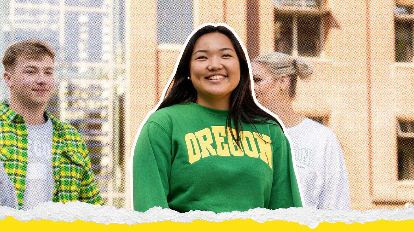A happy student wearing a green Oregon sweatshirt standing outside smiling at the camera. Two other students are talking in the background.