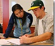 One student sitting at a table with a pencil and paper, with another student standing and looking at the paper