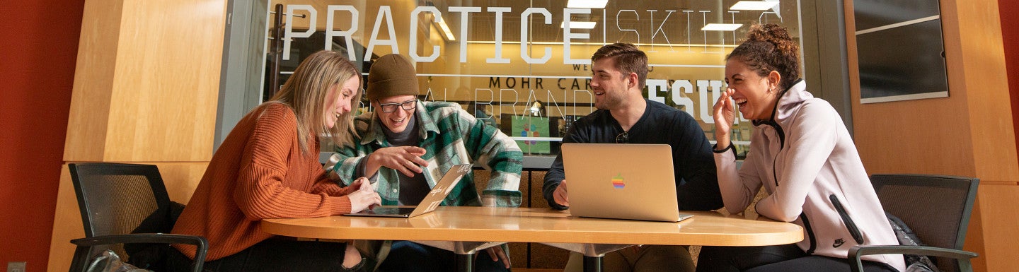 Four students sitting at a table talking and laughing while looking at laptops and working on a project.