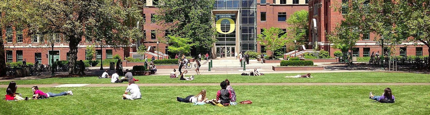 The front of the Lillis Business Complex in the background with grass, tress, and students in the foreground.