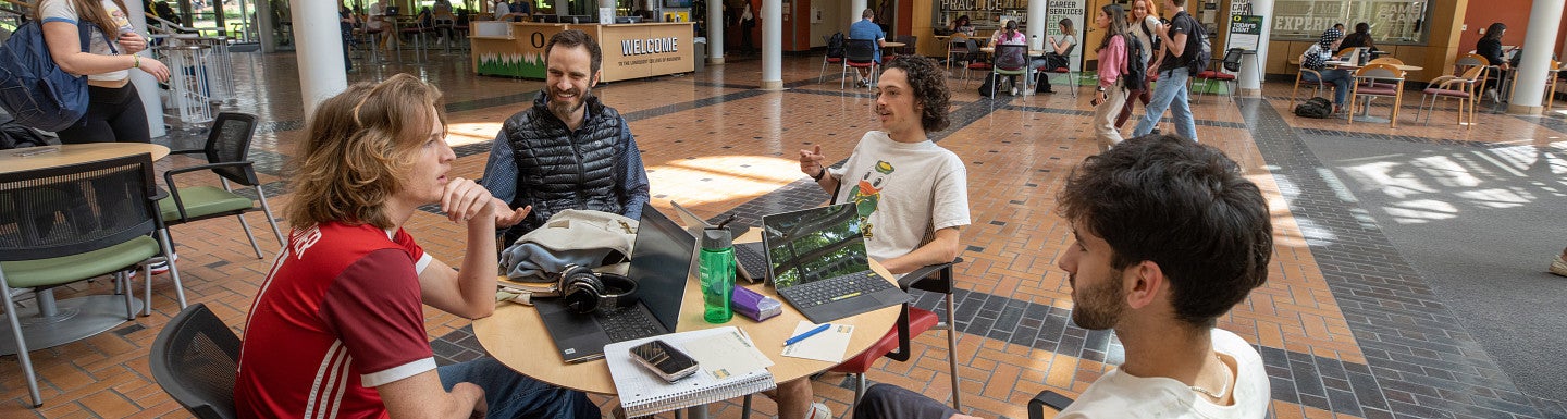 Faculty member and students sitting at table in Lillis atrium having a conversation