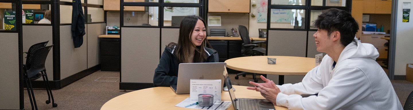 Two students sitting at a round table with laptops, looking at each other, smiling, and talking during a peer advising session