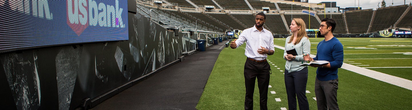 Three students standing on the field of Autzen Stadium looking at a sponsor banner