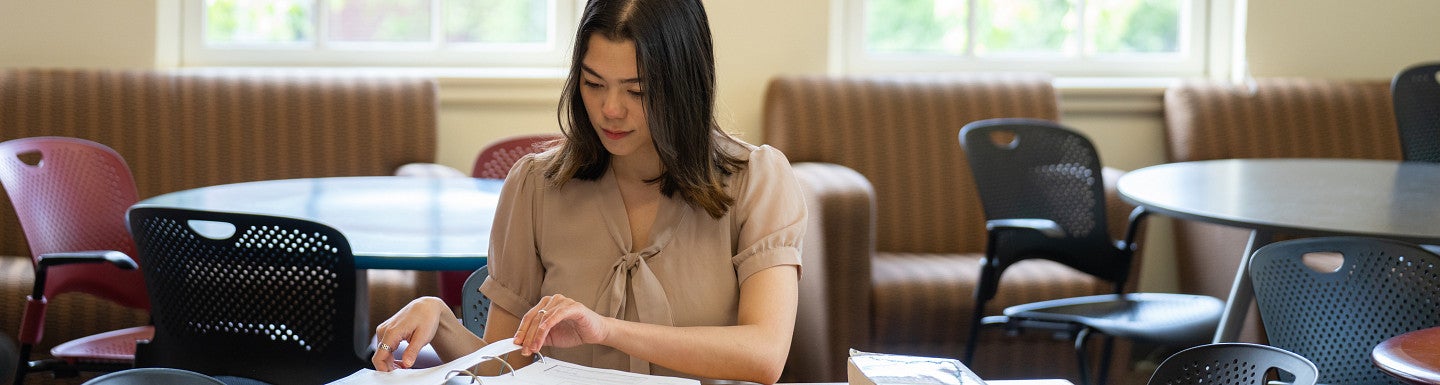 One student sitting at a table with books thumbing through a 3-ring binder 