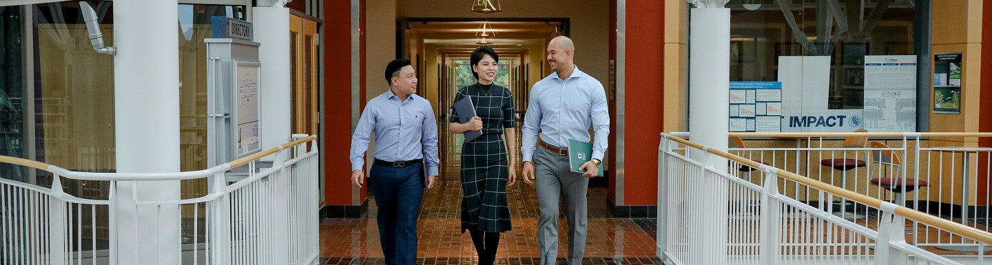 Three students walking side-by-side holding laptops and folders talking with each other.