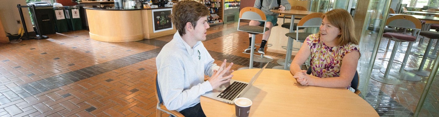 Photo of professor Lanahan meeting with a student while sitting at a round table in the cafe inside the Lillis Business Complex