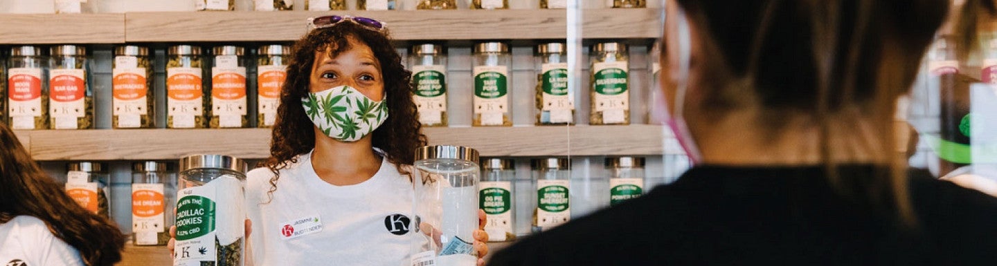 Photo of a women behind the counter and wearing a mask with pot leaves on it at the Kaleafa Cannabis business while helping serve a customer
