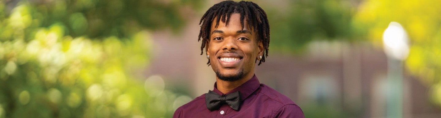 Photo of Jurell Scott smiling at the camera while wearing a maroon shirt with matching bow tie while standing in front of a beautiful bokeh background of colors from campus in spring