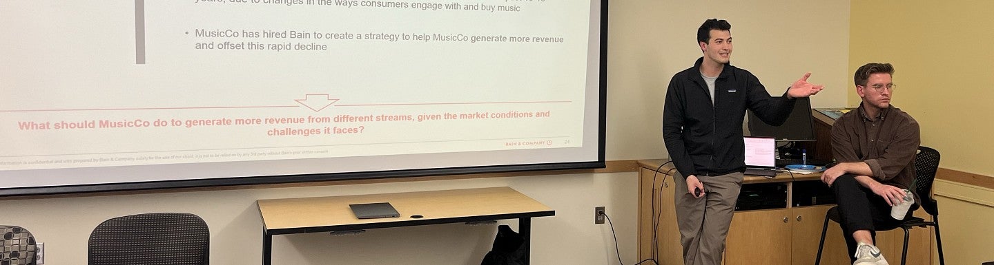 Two guest speakers sitting and standing to the side of a large screen at the front of a classroom, gesturing and speaking to students.