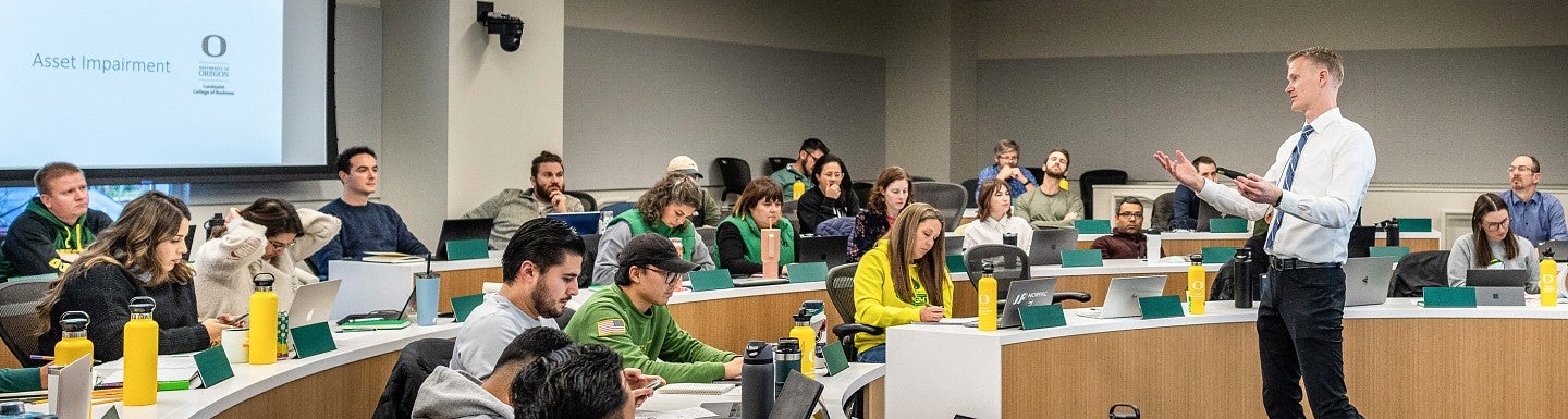 A tiered classroom full of students sitting and looking at the professor who is standing in front of the classroom gesturing and talking..