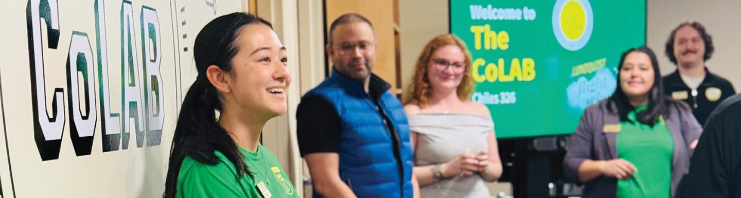 Photo from the opening of the CoLab showing a smiling women student looking sideways toward the audiencein the foreground with CoLAB banner behind her 
