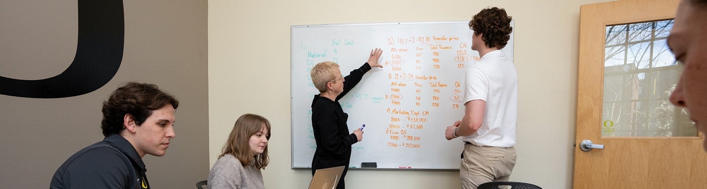 A professor and a student are standing at a whiteboard mid-conversation and pointing at accounting figures. Three other students are sitting at a table on their laptops.
