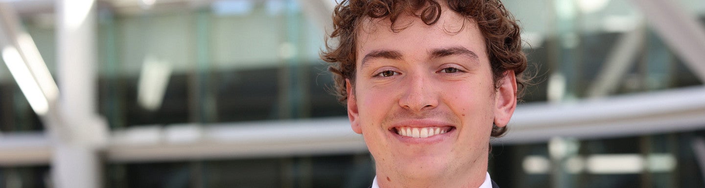 Young man with short curly hair smiles. He is wearing a dark blue suit, white shirt, and dark blue paisley tie.