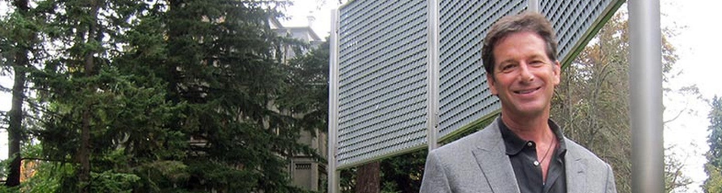 Michael Crook stands in front of the windwall outside the Lillis Business complex in a gray blaze and black shirt smiling at the camera