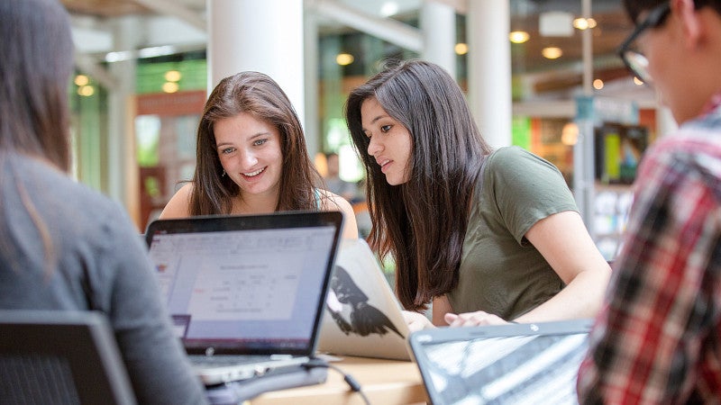 A group of students sitting at a table in the Lillis Business Complex atrium with laptops working on a project.