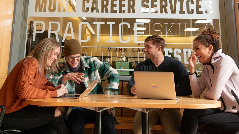 Four students sitting at a table talking and laughing while looking at laptops and working on a project.