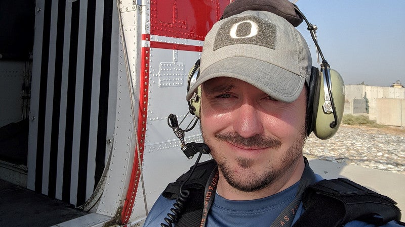 Alum smiling and standing next to a military helicopter wearing headphones with a microphone..