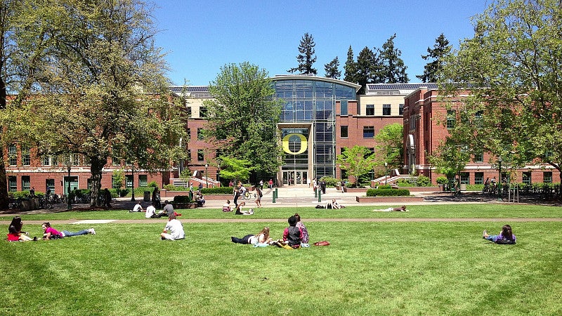 The front of the Lillis Business Complex in the background with grass, tress, and students in the foreground.
