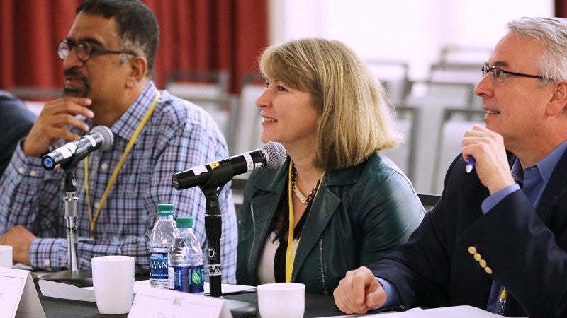 Three people sitting at a table with microphones smiling and looking at a presenter.