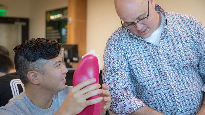 Faculty member talking to and standing next to a student who is sitting at a table, holding a shoe mold, and working on a project.