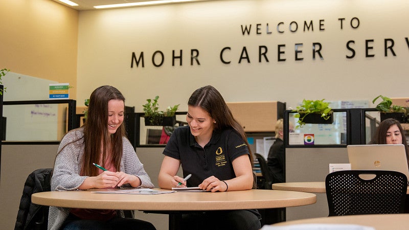 Two students sitting at a table smiling and looking at paperwork.