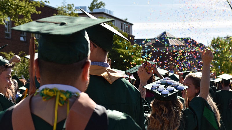 A crowd of students dressed in commencement regalia throwing confetti into the air.