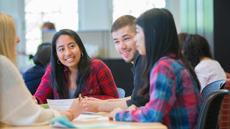 Student sitting at a table, talking and looking at papers on the table.