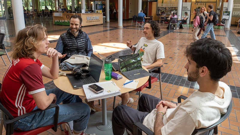 Faculty member and students sitting at table in Lillis atrium having a conversation