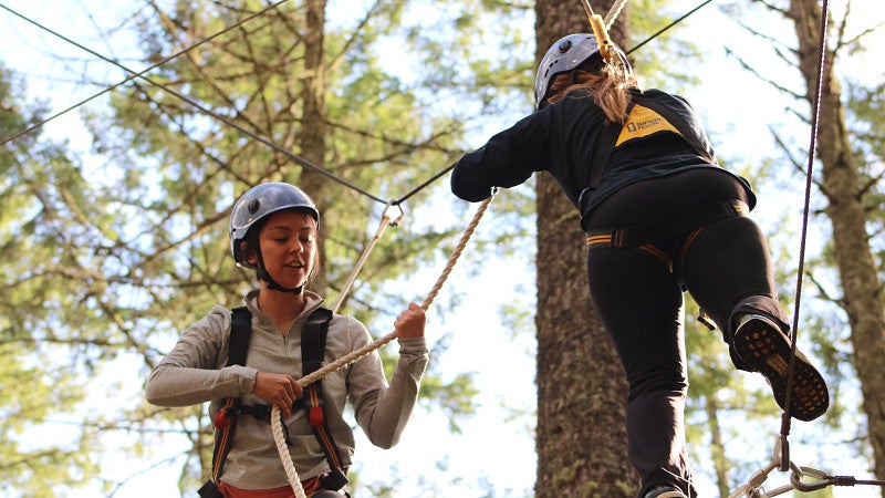 Two students wearing safety helmets and harnesses walking a ropes course high in the trees