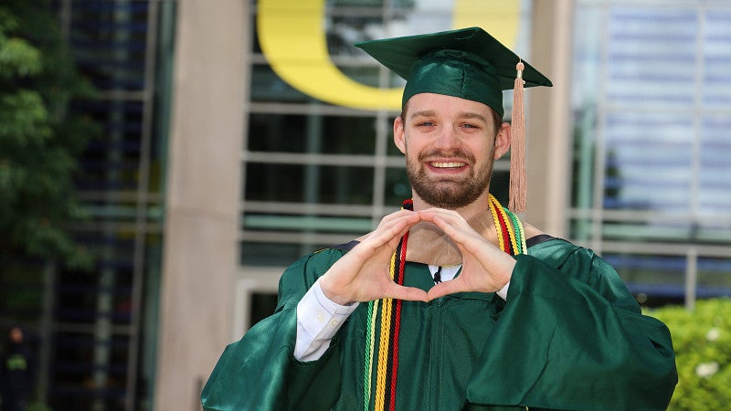Student outside looking at the camera wearing commencement regalia and holding their hands in the shape of an O