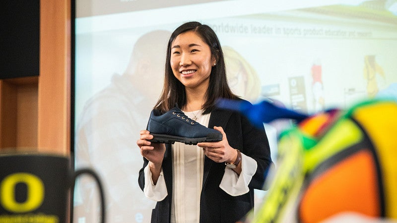 Student at front of the class standing in front of a large screen holding up an athletic shoe