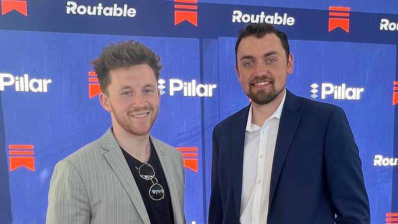 Two alumni standing in front of a media backdrop looking at the camera and smiling