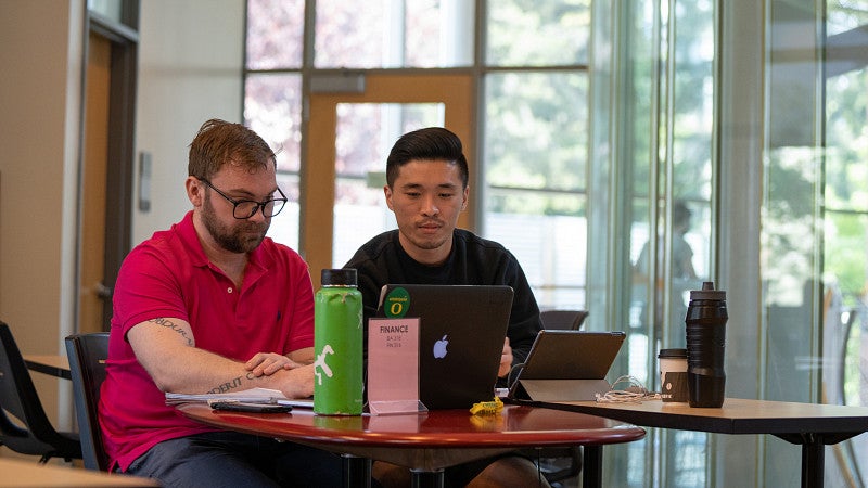 Two students in the distance sitting at a round table with laptops during a tutoring session