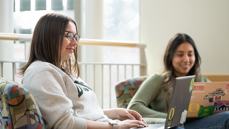 Two students sitting in soft chairs smiling and working on laptops