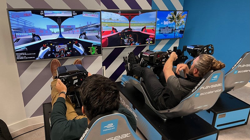 Two MBA students are seated in arcade-style racing seats in front of wall-mounted monitors which display the driver's interior view of a car.