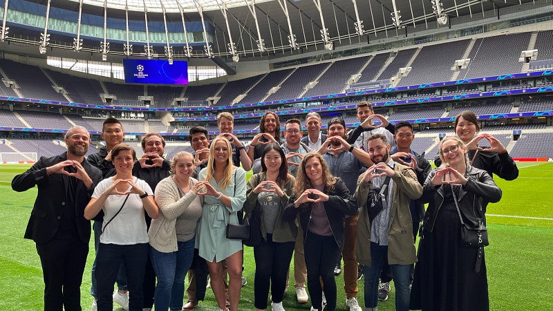 A group of MBA students pose for a photo in a stadium, making the University of Oregon "O" symbol with their hands. They are standing on the stadium turf. The empty stands and stadium awning can be seen behind them.