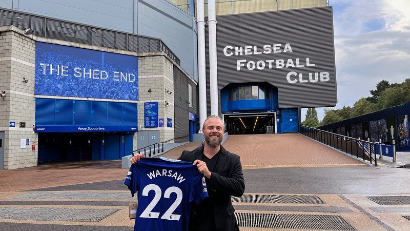 Warsaw Sports Marketing Center program manager Robert Greisinger smiles and holds up a blue number 22 jersey with the word "Warsaw" in place of a player's name. Behind Greisinger is a large building; on the right side the words "Chelsea Football Club" are