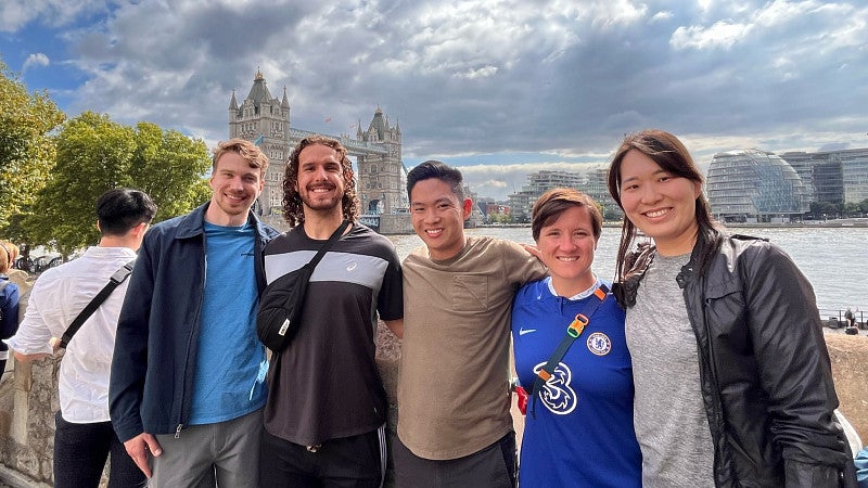 A group of five MBA students smile and pose for a photo in front of the river Thames. Behind them are the Tower Bridge and London's Old City Hall.