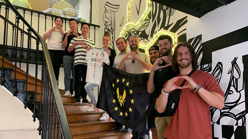 Eight MBA students and a staff member are standing along a staircase from bottom to top, making the University of Oregon "O" symbol with their hands. Several of them are holding a up black flag with a yellow sigil. Another student is holding up a white shirt with text on it.