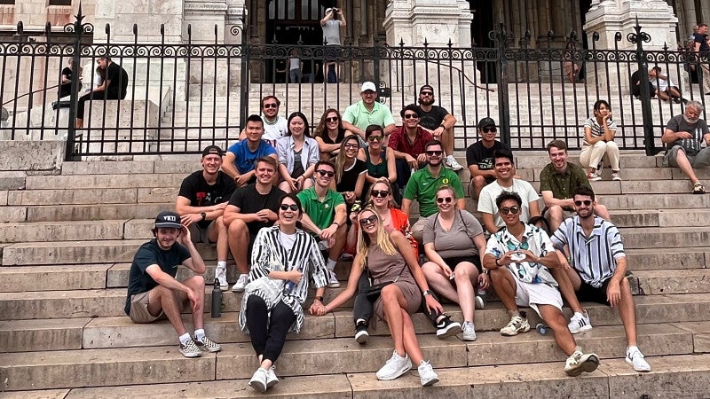 A group of MBA students are seated on the steps of a cathedral in Paris. Behind them, between the steps and the cathedral entrance, is a black iron fence. Statues and other designs can be seen on the exterior of the cathedral.