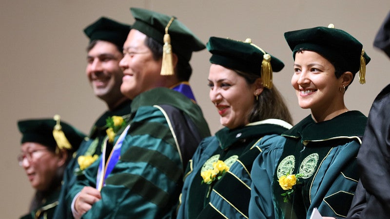 Five students standing and smiling, wearing doctoral regalia at a commencement ceremony.