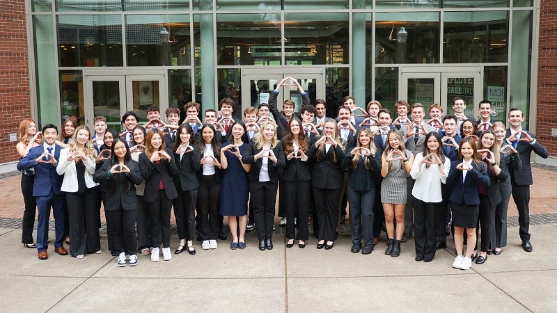 A group photo of business honors students standing in front of the Lillis Business Complex holding their hands in the shape of an O.