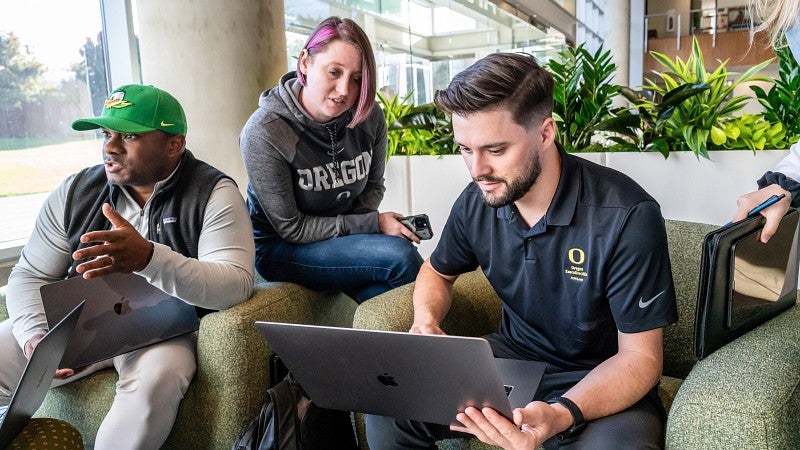 Three students sitting on soft seating looking at a laptop and talking with each other.