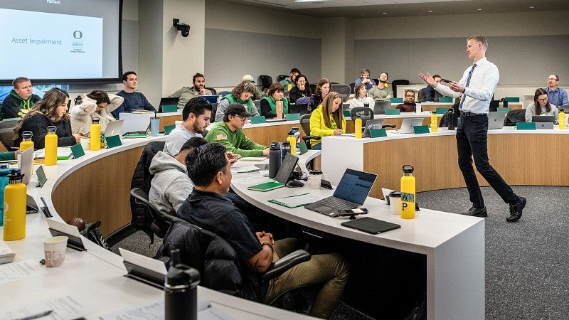 A tiered classroom full of students sitting and looking at the professor who is standing in front of the classroom gesturing and talking..