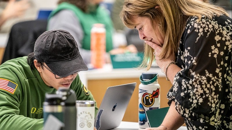 One student sitting down at a desk looking at a computer while another student is standing on the other side of the desk leaning towards the first student.