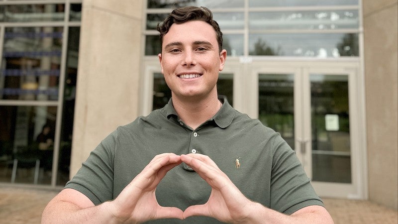 Alex La China standing in front of the Lillis Business Complex smiling and throwing the O.