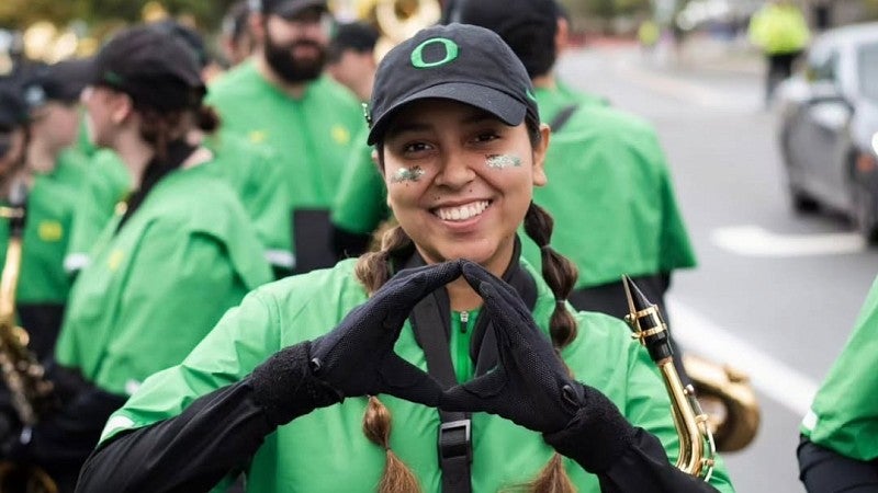 Victoria Castillo is standing outside in a crowd dressed in Duck gear and holding a saxophone while smiling at the camera and throwing the O in excitement.
